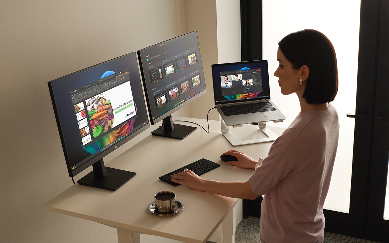 Fotografi af en kvinde, der bruger en Copilot+ PC bærbar computer på en riser tilsluttet to skærme, hvor hun arbejder på en PowerPoint præsentation.