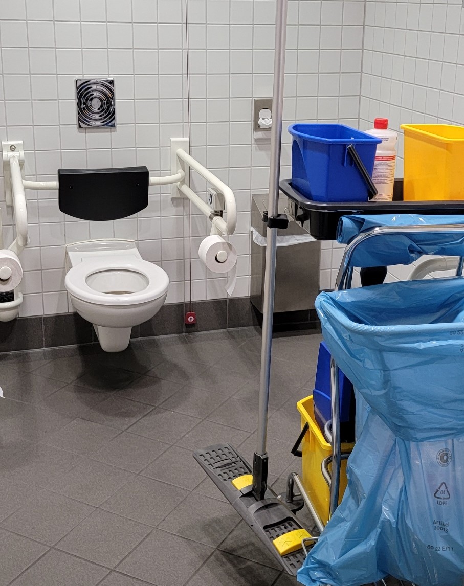 A cleaning cart sits next to a toilet in a bathroom for people with disabilities.