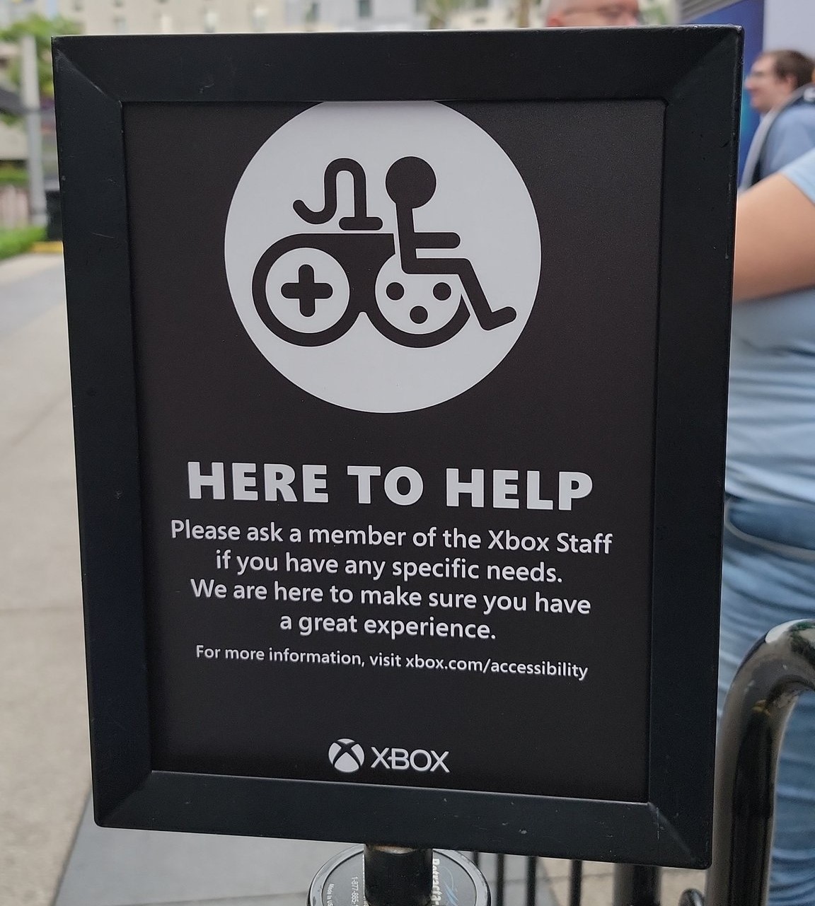A black sign with white text which reads “Here to Help."; A gaming accessibility logo appears on it. Text reads: "Please ask a member of the Xbox Staff if you have any specific needs. We are here to make sure you have a great experience. For more information, visit xbox.com/accessibility."