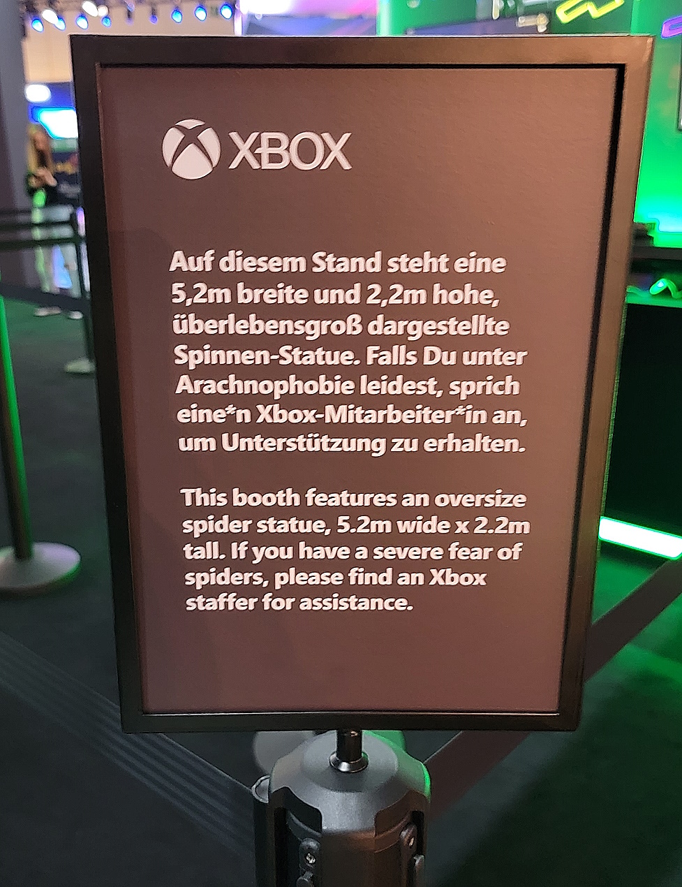 A black sign with white text which reads in both English and German, "This booth features an oversize spider statue, 5.2m wide x 2.2m tall. If you have a severe fear of spiders, please find an Xbox staffer for assistance.