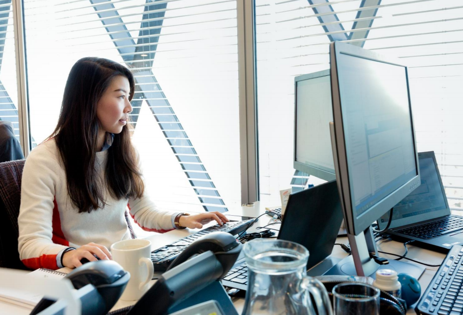 Eine Frau sitzt an einem Bürotisch vor einem Laptop und einem Monitor.
