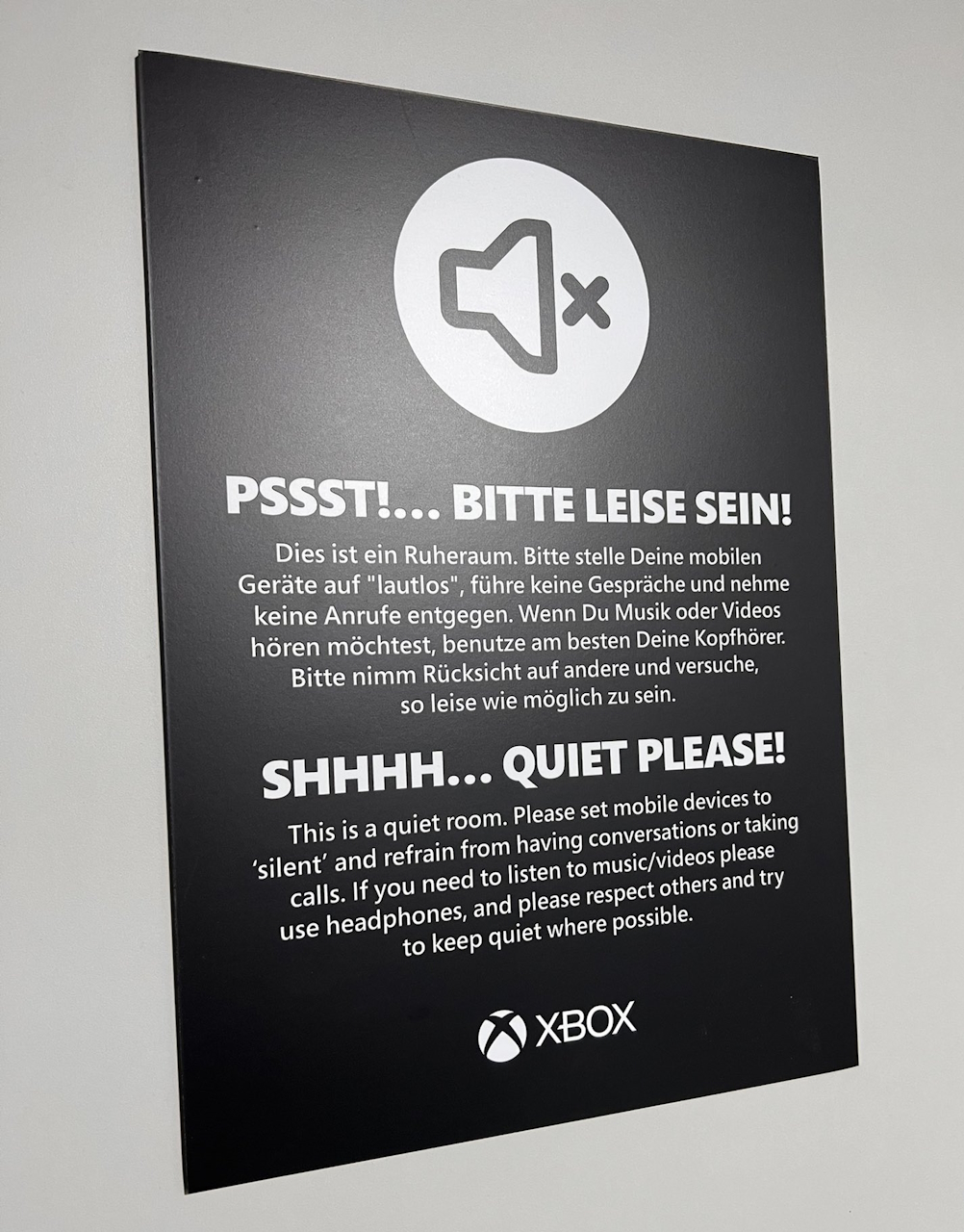 A black sign with white text which reads in English and German, "Shhhh... Quiet Please! This is a quiet room. Please set mobile devices to silent and refrain from having conversations or taking calls. If you need to listen to music/videos please use headphones, and please respect others and try to keep quiet where possible."