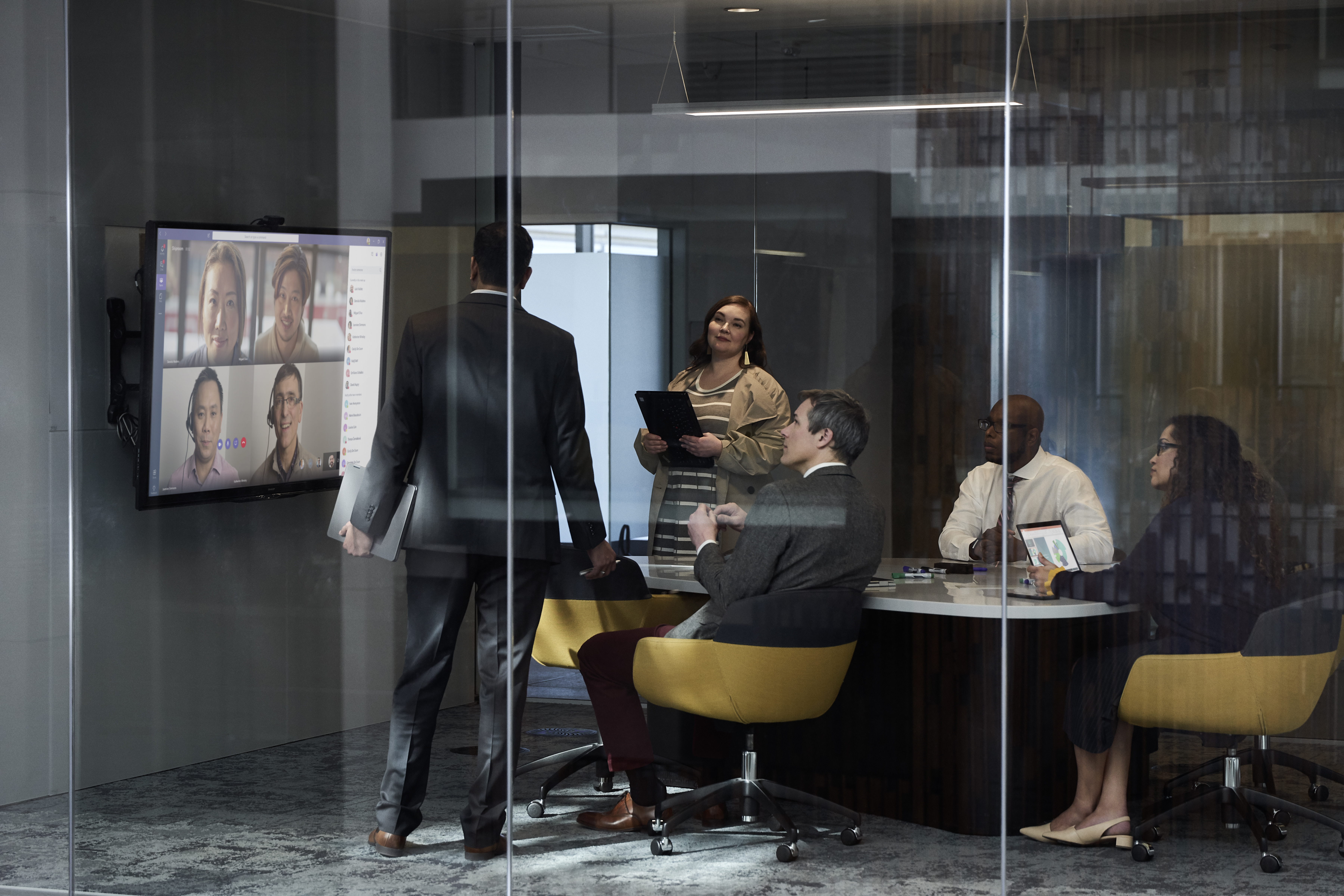 A group of office workers in a meeting discuss images on a large scree.