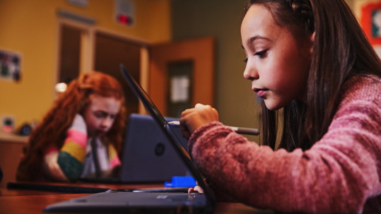 Two young girls in a classroom setting, one using a laptop and one using a tablet and stylus.