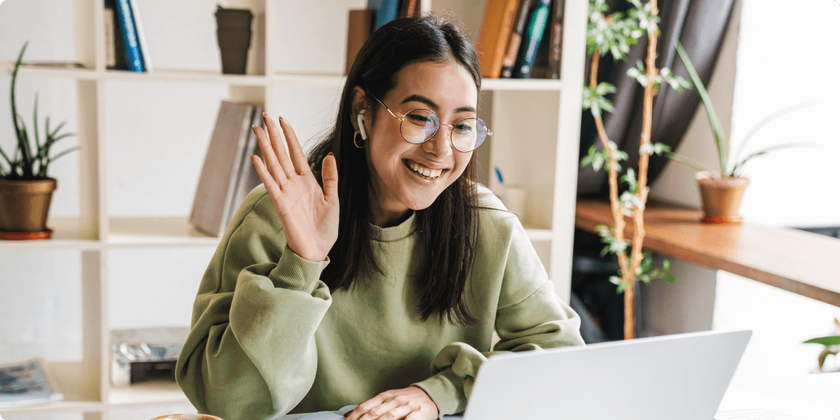 A woman on her laptop smiling and waving to the screen. She is in a home office setting.