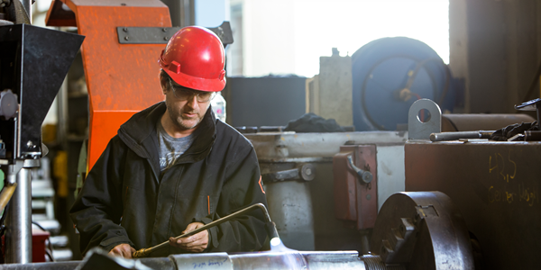 A man with a hard red hat and a safety jacket is using a blow torch on manufacturing equipment.