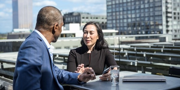 Male and female corporate office workers in suits sitting on rooftop deck. The woman is holding a phone and there is a closed laptop on the table.