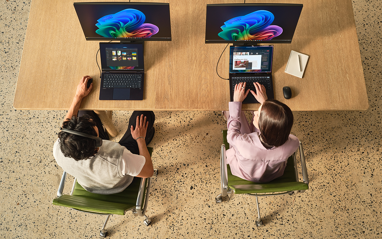 Overhead photograph of two employees using Windows laptops and external monitors at a shared workspace.