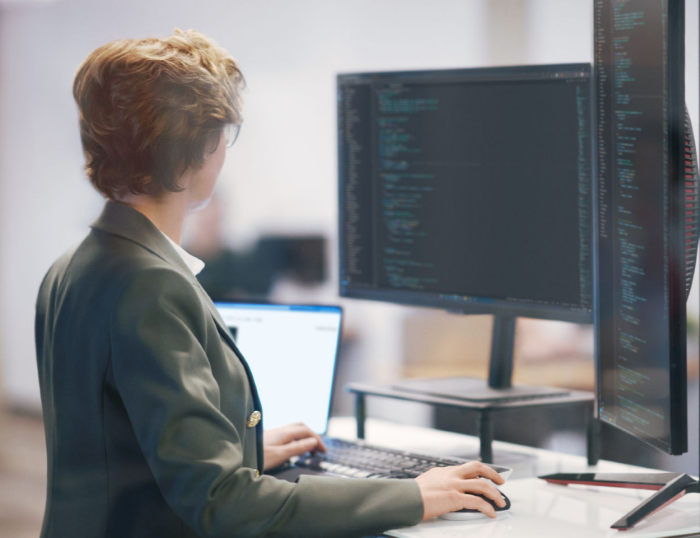 A photograph of a person working at a standing desk with three monitors displaying code.