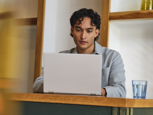Photograph of a person using a laptop at a wooden desk with a glass of water nearby.