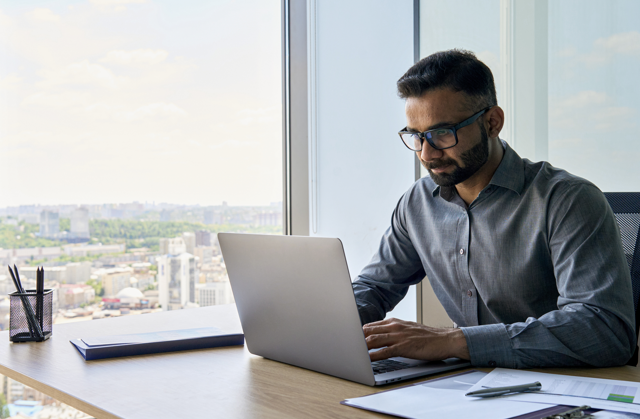 Photograph of executive sitting at desk using laptop in an office with a panoramic view.
