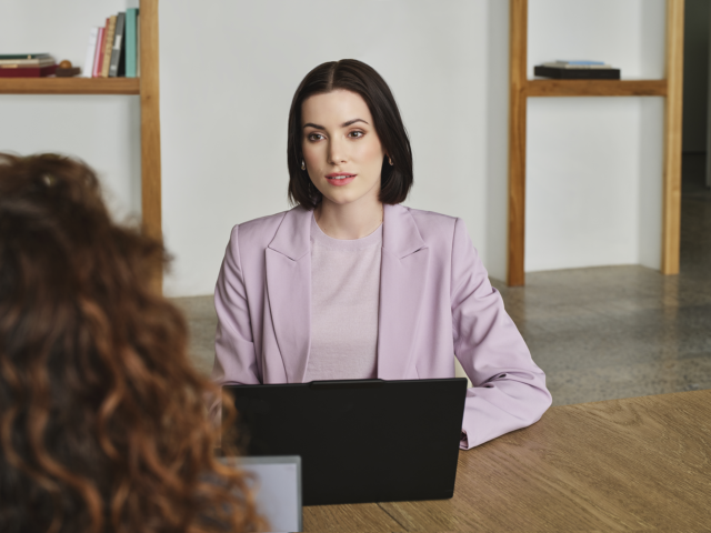 Photograph of two people talking in an office setting, with one person seated at a desk using a laptop.