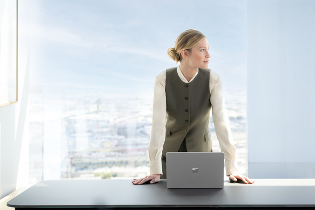 A photograph of a business professional standing at their desk with their Surface device in front of a large window.
