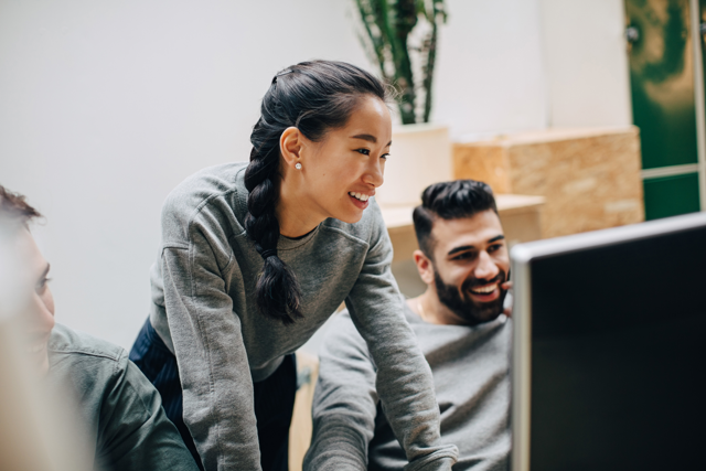 A photograph of two employees collaborating at a desktop computer, one seated and the other standing beside them in an office.