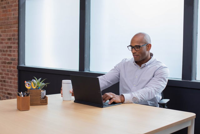A photograph of an employee smiling while working on a laptop at a desk in an office building.