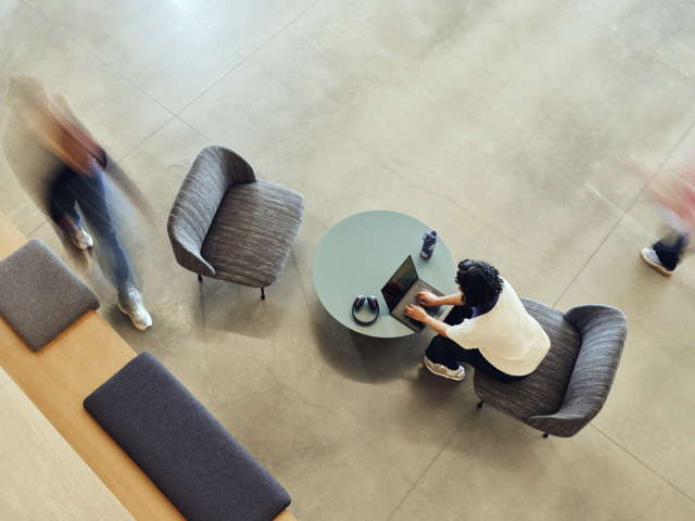 A photograph showing an arial view of a remote worker using a laptop while seated in a building lobby.
