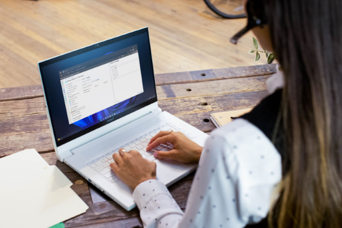 A photograph of an IT professional using a white laptop at a wooden table with documents and a notebook.