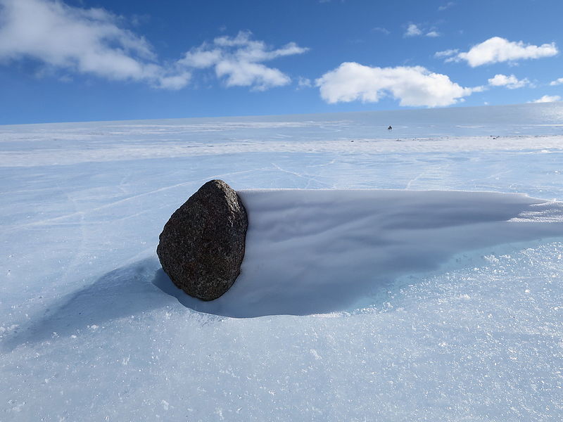 Photo of a black rock sitting in an indentation on an ice field.