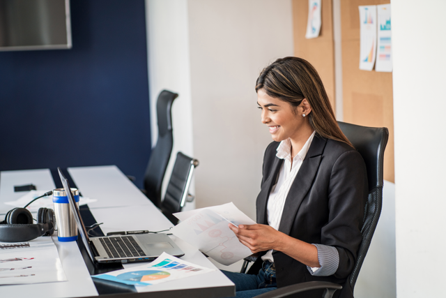 A woman smiling while working on a laptop in an office environment. They are holding datasheets with a bar graph printed on the paper.