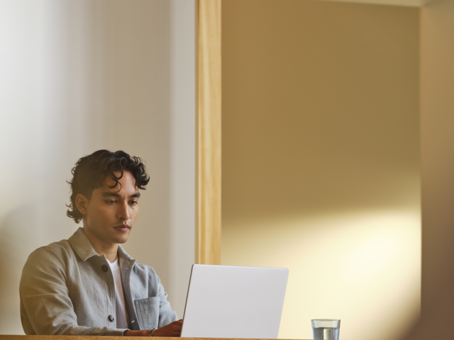 A photograph of a young man working at a desk in an office on a laptop.