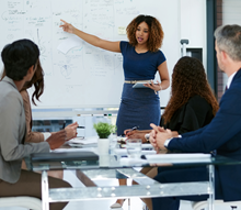 A person points to a whiteboard as other people listen.