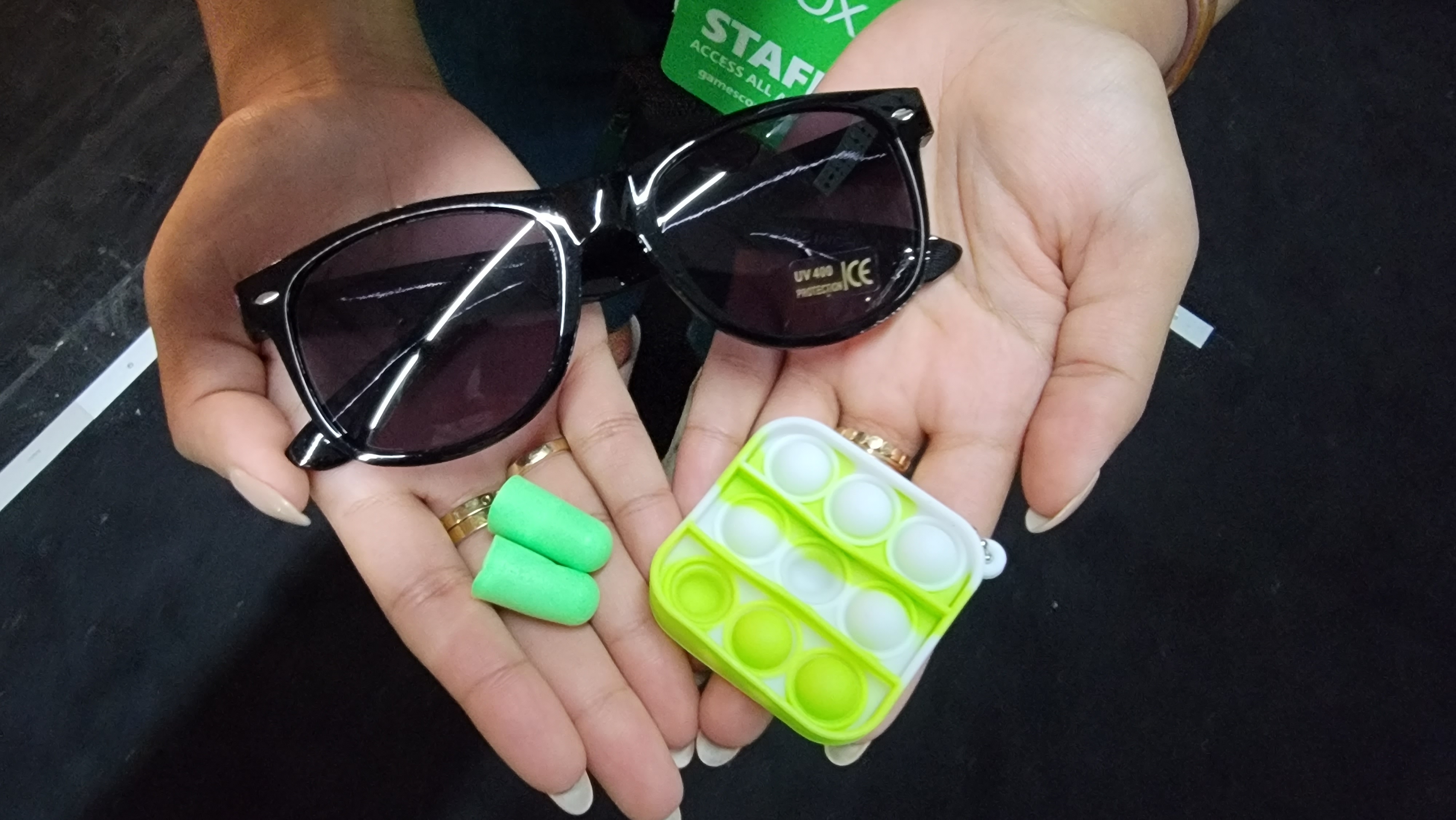 A pair of hands holds dark glasses, a fidget toy, and earplugs.