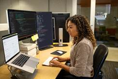 Une femme, assise à son bureau, effectue des tâches de programmation sur un ordinateur portable connecté à plusieurs moniteurs d’ordinateur de grande taille.