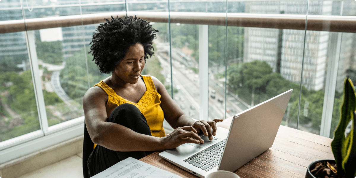 Une femme travaillant sur son ordinateur portable, sur son balcon. Derrière elle, on aperçoit des arbres et des tours d’habitation.