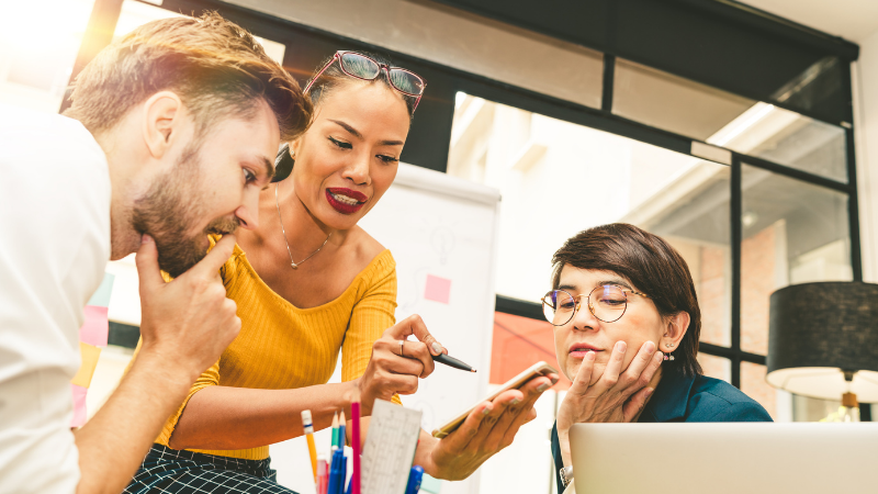 Photograph of a group of three teachers engage in a project brainstorm discussion at their school, using a smartphone.