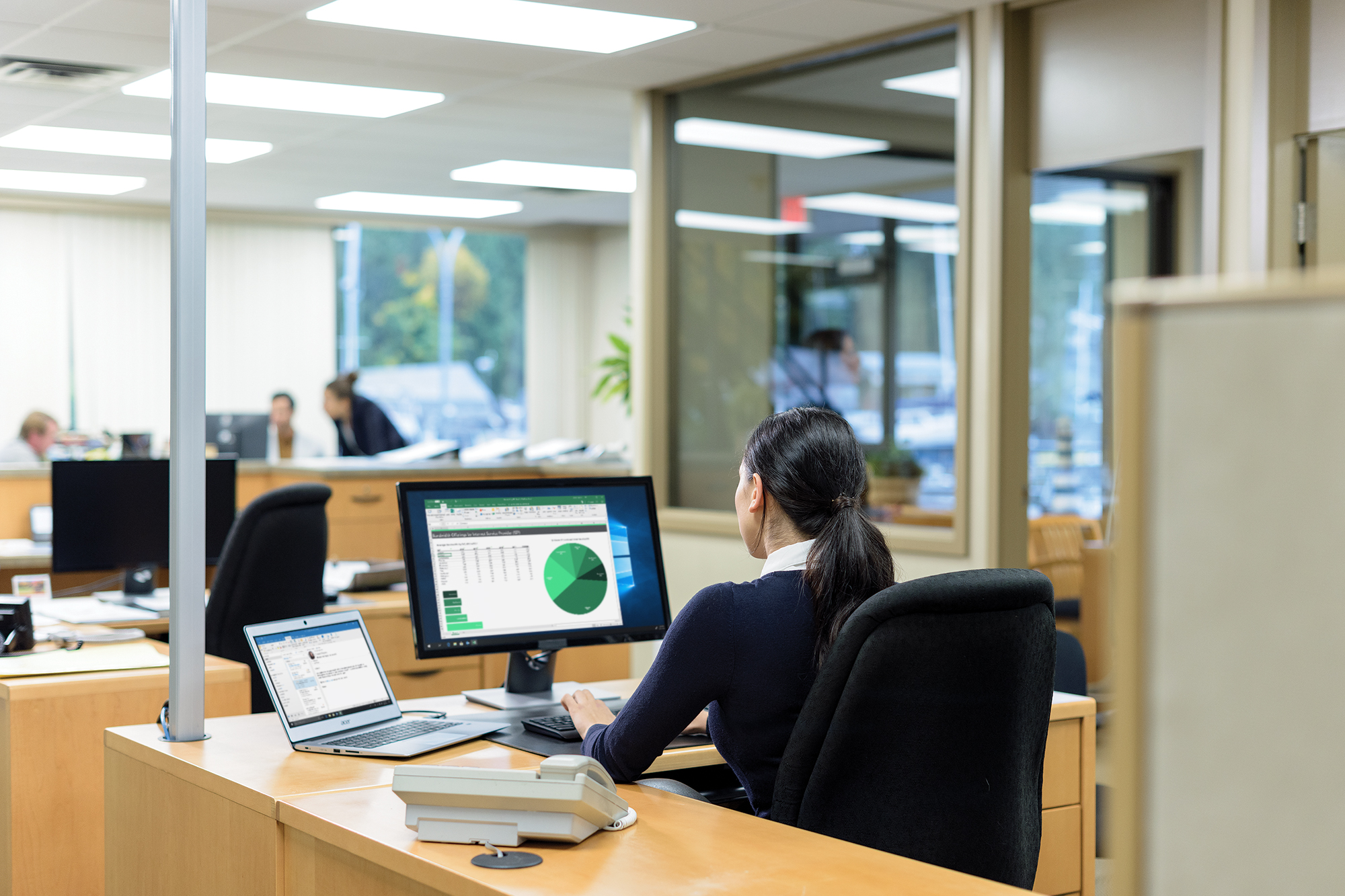 Een kantoormedewerker bekijkt een grafiek en tabellen op een scherm, terwijl anderen op de achtergrond vergaderen.