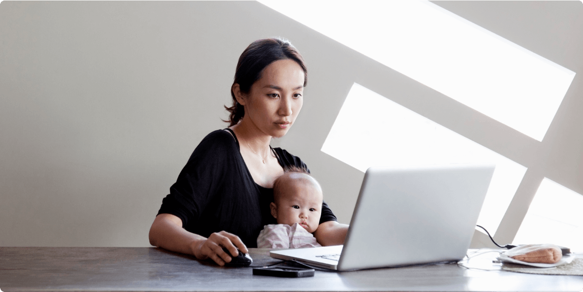 Een moeder met haar peuter op haar schoot, die op een laptop werkt. Zonlicht stroomt achter haar aan.