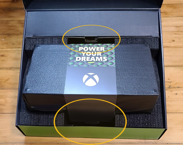 Uma imagem do empacotamento do Xbox Series X, aberta e com um console dentro. Dois círculos laranjas realçam uma seção dentro da embalagem e na frente da embalagem.