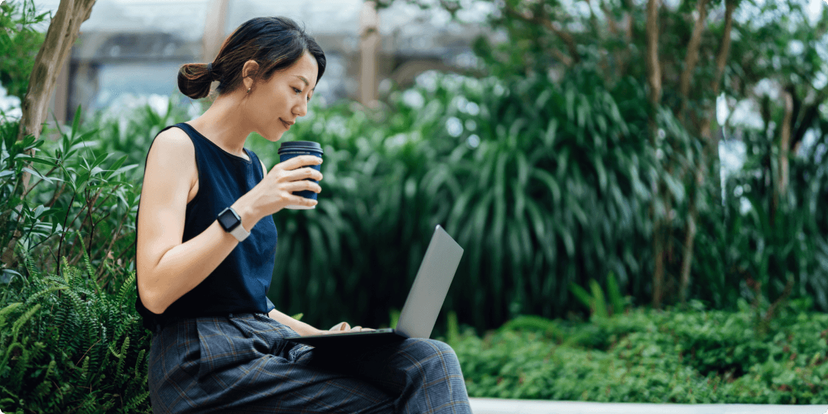 Uma mulher em seu laptop do lado de fora com uma xícara de café. Ela está em torno de uma vegetação exuberante.