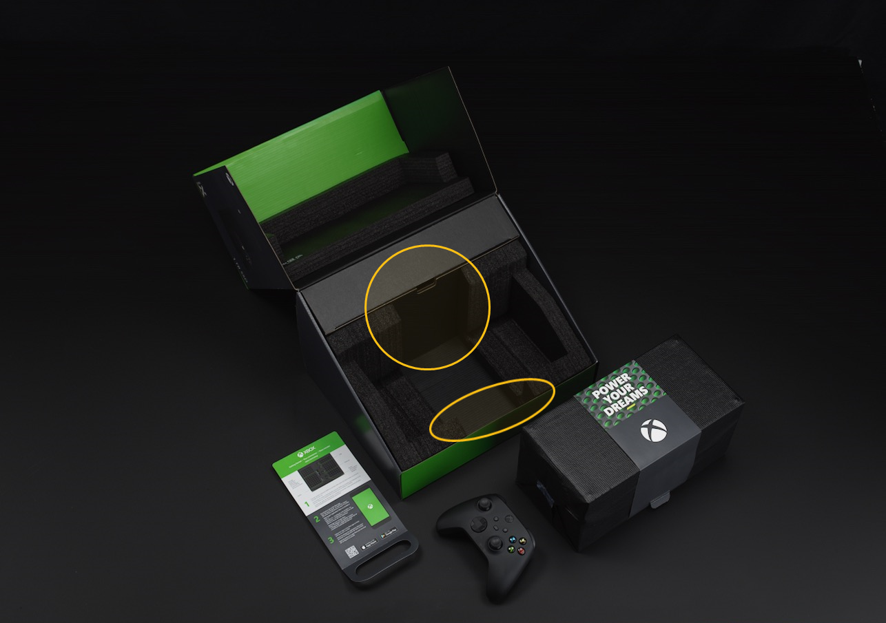 Uma imagem da embalagem do Xbox Series X, aberta e vazia. Dois círculos laranja destacam uma secção no interior da embalagem e na parte da frente da embalagem.