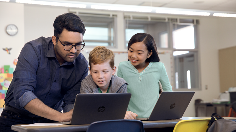 Foto de um professor apoiando dois alunos que estão usando laptops para concluir o trabalho.