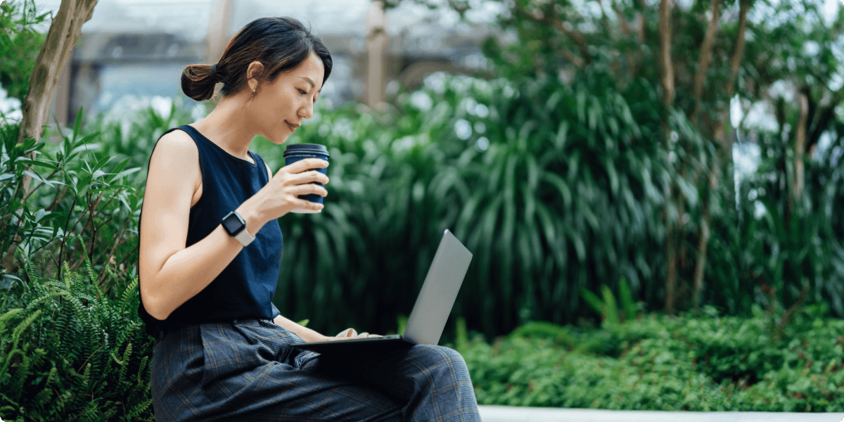 O femeie de pe laptopul ei afară cu o ceașcă de cafea. Ea este în jurul verdeață luxuriantă.