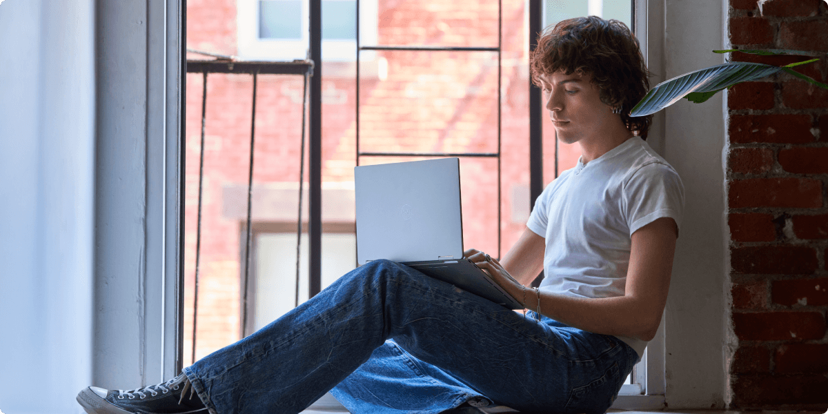 Genç bir adam gündelik olarak dizüstü bilgisayarıyla pencere kenarında oturuyor.