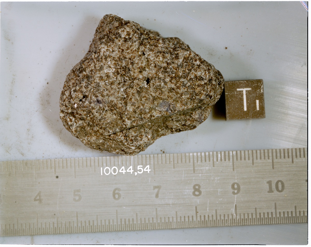 A photo of a rock on a flat surface. The rock is positioned next to a ruler. The ruler shows the approximate size for the rock as 4 inches. Another numerical measurement on the ruler reads 1 0 0 4 4 comma 5 4. A small cube next to the rock has the label T one.