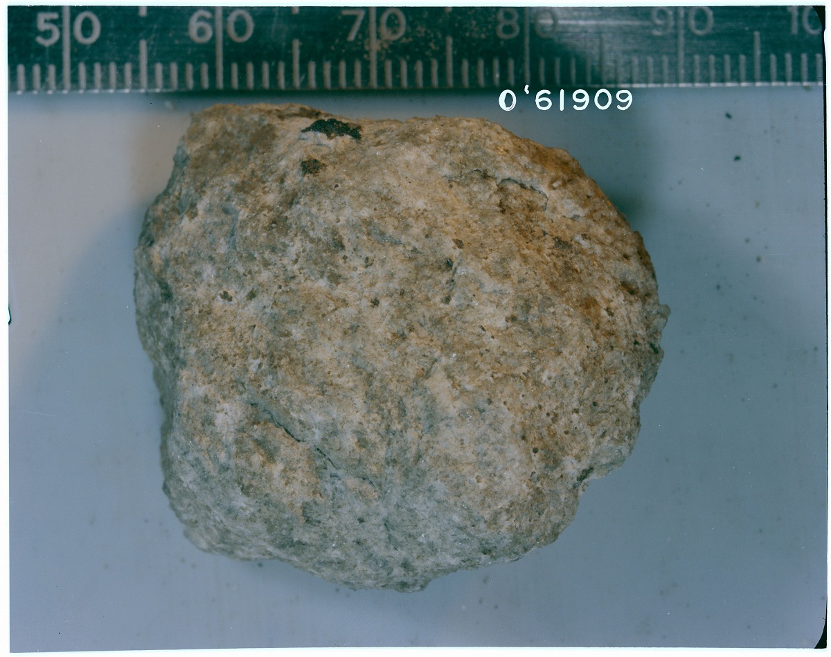 A photo of a piece of highland rock on a flat surface. The rock is positioned next to a ruler. The ruler shows the approximate size for the rock as three and one-half inches. Another numerical measurement on the ruler reads zero apostrophe 6 1 9 0 9.