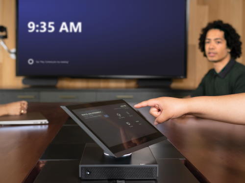 A user taps a Teams Rooms console, with a display in the background.